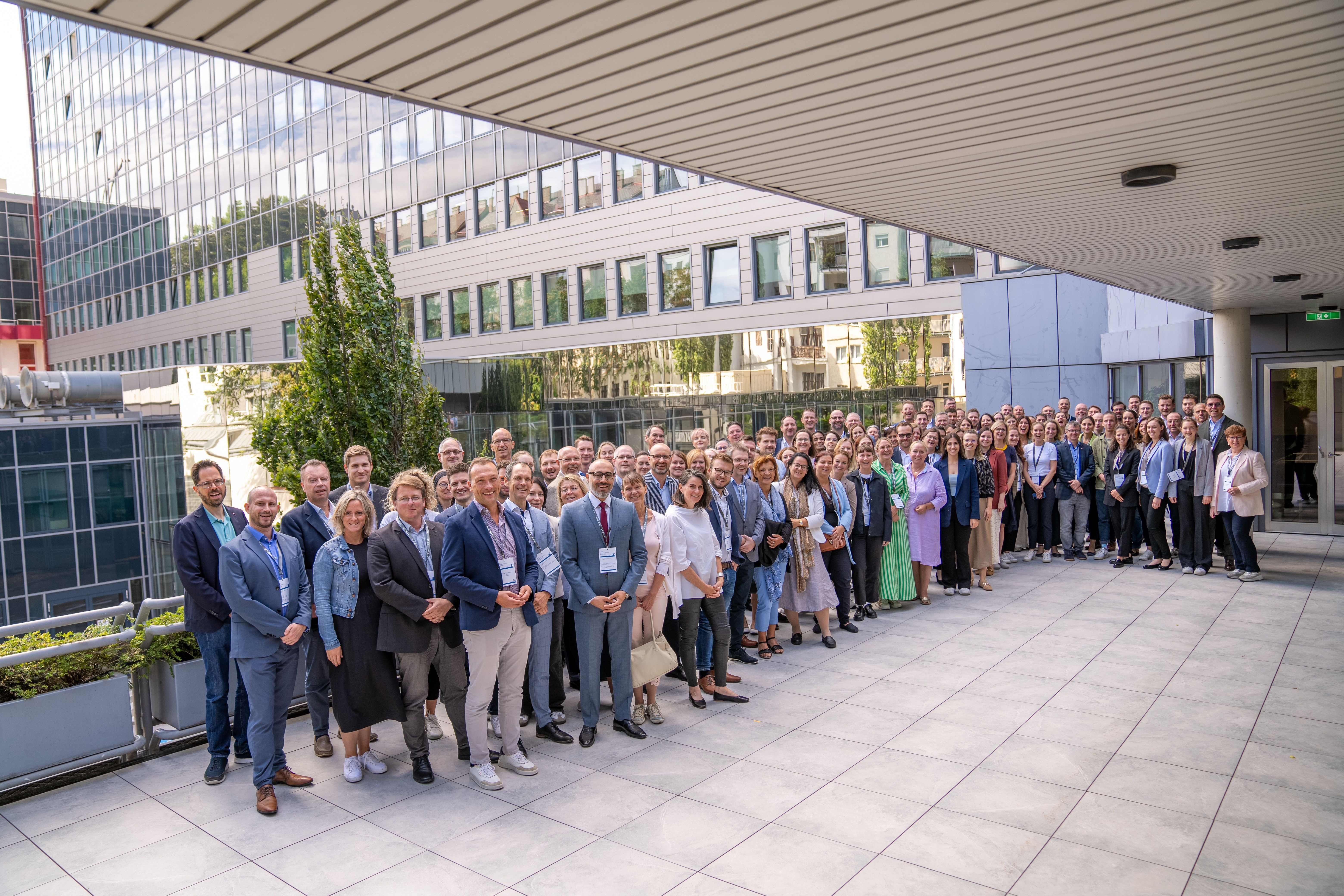 Bild: Gruppenfoto vieler verschiedener Personen, die im Außenbereich eines Gebäudes stehen. Die Personen tragen unter anderem Sakkos und Ausweisschilder um den Hals. Im Hintergrund ist die Fassade eines Gebäudes